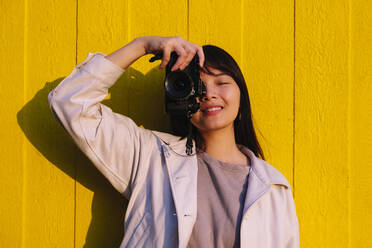 Smiling young woman photographing in front of yellow wall - ASGF02662