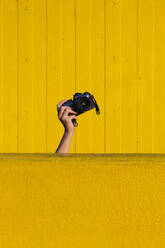 Hand of woman holding camera behind yellow wall - ASGF02663