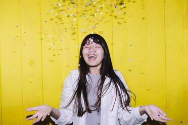 Cheerful young woman standing under confetti in front of yellow wall - ASGF02666