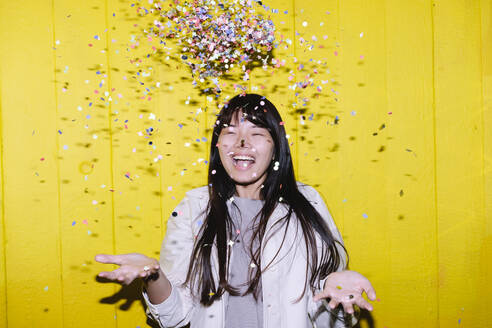Fröhliche junge Frau wirft Konfetti vor einer gelben Wand - ASGF02667