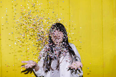 Happy woman throwing multi colored confetti in front of yellow wall - ASGF02668