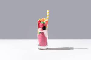 Glass with bunch of sweet gummy candies and pink sugar placed near striped straw on grey and white background - ADSF36200