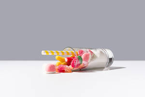 Fallen Glas mit Bündel von süßen Gummibonbons und weißen Zucker in der Nähe von gestreiften Stroh auf grauem und weißem Hintergrund platziert - ADSF36204