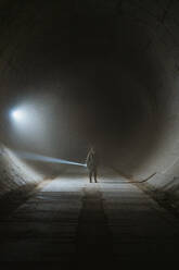 Person with flashlight illuminating darkness and exploring grungy underground tunnel with concrete walls during apocalypse - ADSF36216