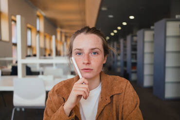 Junge Studentin, die ihr Gesicht mit einem Stift berührt und in die Kamera schaut, während sie Hausaufgaben auf einem unscharfen Hintergrund einer Bibliothek macht - ADSF36221