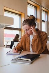 Junge Frau in Freizeitkleidung mit Haarknoten, die den Kopf berühren und in einen Planer schreiben, während sie am Tisch sitzt und in der Universitätsbibliothek studiert - ADSF36225