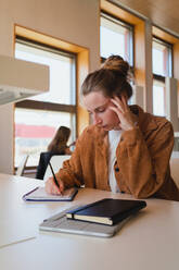 Young woman in casual clothes with hair bun touching head and writing in planner while sitting at table and studying in university library - ADSF36225