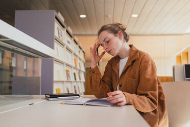 Junge Frau in Freizeitkleidung mit Haarknoten, die den Kopf berühren und in einen Planer schreiben, während sie am Tisch sitzt und in der Universitätsbibliothek studiert - ADSF36232
