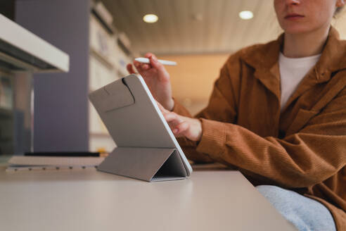 Unrecognizable woman with stylus making notes on tablet while doing homework assignment in library in daytime - ADSF36234