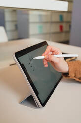 Unrecognizable woman with stylus making notes on tablet while doing homework assignment in library in daytime - ADSF36236