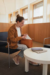Junge Studentin in Freizeitkleidung mit Haarknoten, die Schreibwaren aus der Tasche nimmt, während sie auf einem Stuhl in der Nähe des Tisches sitzt und sich auf das Studium in der Universitätsbibliothek vorbereitet - ADSF36239