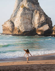 Back view female traveler walking on sandy coast with rough rocky cliffs near sea at sunset time on sunny summer evening - ADSF36243