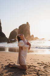 Full body side view of female traveler sitting on sandy coast near waving waving with rocky cliffs at sunset time - ADSF36245