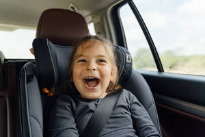 Fröhliches kleines Mädchen mit braunen Haaren in Freizeitkleidung, das lächelt und wegschaut, während es im Autositz sitzt, während einer Autofahrt auf dem Lande - ADSF36249