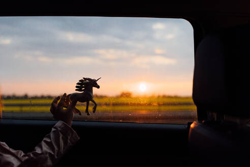 Crop anonymous kid playing with horse toy near window of riding car in countryside during sunset - ADSF36253