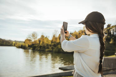 Frau mit Mütze fotografiert See durch Smartphone an sonnigem Tag - ACPF01444