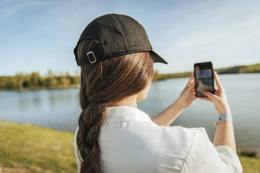 Junge Frau mit Mütze fotografiert See mit dem Smartphone - ACPF01446
