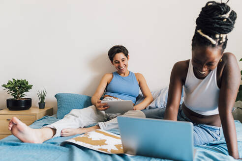 Happy woman using laptop with girlfriend in background on bed at home - MEUF07857