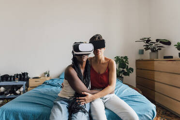 Lesbisches Paar mit VR-Brille sitzt zu Hause auf dem Bett - MEUF07950