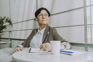 Geschäftsfrau mit Brille sitzt mit Tagebuch und Einwegbecher in einem Café - OIPF02305
