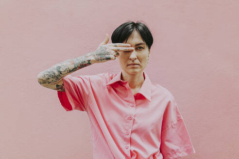 Tätowierte Frau mit kurzen Haaren, die das Auge mit der Hand vor einer rosafarbenen Wand bedeckt - OIPF02319