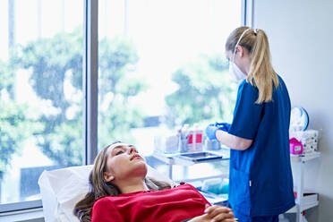 Junge Frau mit grauen Augen liegt auf dem Tisch und lächelt während eines Termins in einer Schönheitsklinik am Tag mit einer Kosmetikerin - ADSF36256