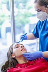 Esthetician in uniform and face mask injecting collagen into forehead of female client near window in office of beauty clinic. - ADSF36257