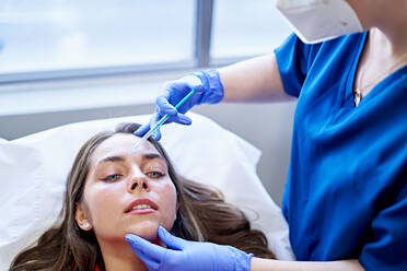 Cropped unrecognizable esthetician in uniform and face mask injecting collagen into forehead of female client near window in office of beauty clinic. - ADSF36258