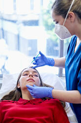 Esthetician in uniform and face mask injecting collagen into forehead of female client near window in office of beauty clinic. - ADSF36259