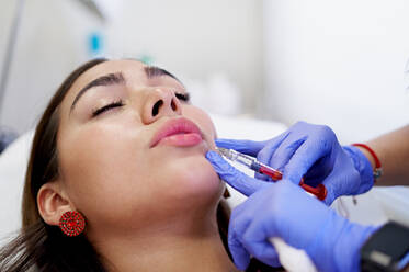 From above crop unrecognizable esthetician in latex gloves injecting collagen into lips of relaxed female client during appointment in beauty clinic - ADSF36265