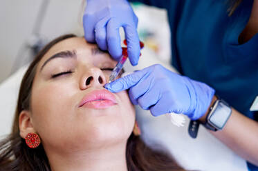 From above crop unrecognizable esthetician in latex gloves injecting collagen into lips of relaxed female client during appointment in beauty clinic - ADSF36266