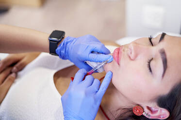 From above crop unrecognizable esthetician in latex gloves injecting collagen into lips of relaxed female client during appointment in beauty clinic - ADSF36267