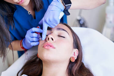 From above crop unrecognizable esthetician in latex gloves injecting collagen into lips of relaxed female client during appointment in beauty clinic - ADSF36268