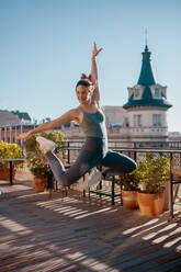 Aktive lächelnde Frau in Sportkleidung, die mit erhobenem Arm springt, während sie während des Trainings auf der Terrasse an einem Sommertag Übungen macht - ADSF36279