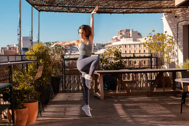 Ganzer Körper einer aktiven Frau in Sportkleidung bei einer Übung mit angehobenem Bein auf der Terrasse eines Gebäudes während des Trainings an einem Sommertag - ADSF36282