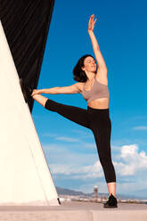 Full body of sporty female in activewear with eyes closed and raised arm doing split and stretching leg on wall during outdoor training against blue sky on summer day - ADSF36289