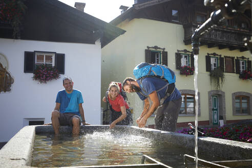 Fröhlicher Wanderer sitzt neben verspielten Freunden, die am Brunnen im Dorf Mutters, Tirol, Österreich, Wasser spritzen - CVF02174