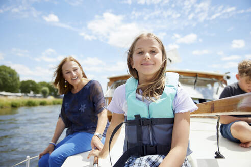 Lächelndes Mädchen mit Schwimmweste auf dem Bootsdeck sitzend mit Familie im Urlaub - RHF02600
