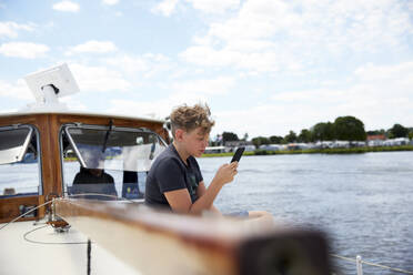 Boy using smart phone sitting on boat deck - RHF02617