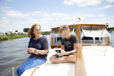 Smiling grandmother with grandson using smart phone sitting cross-legged on boat deck - RHF02625
