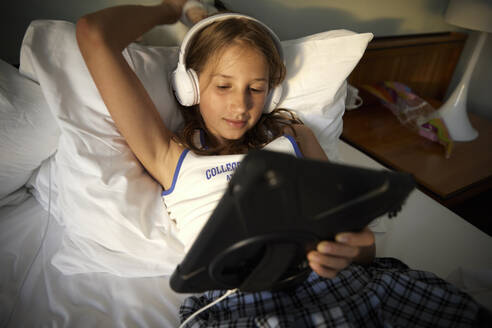 Mädchen mit erhobener Hand und Kopfhörern, das einen Tablet-PC benutzt und sich auf einem Bett in einem Motorboot entspannt - RHF02648