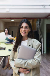 Porträt einer lächelnden brünetten Geschäftsfrau mit Laptop im Büro - PNAF04365