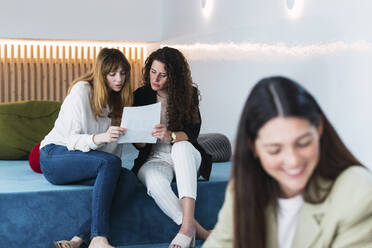 Zwei Geschäftsfrauen lesen ein Dokument in einer Bürolounge mit einem Kollegen im Vordergrund - PNAF04448