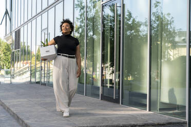 Smiling young businesswoman with box walking on footpath - VPIF06944