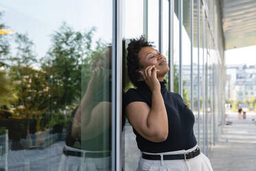 Lächelnde Geschäftsfrau mit geschlossenen Augen, die mit einem Smartphone spricht und sich an eine Glaswand lehnt - VPIF06950