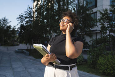 Smiling young businesswoman with eyes closed holding laptop talking on mobile phone - VPIF06959