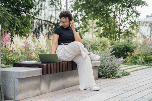 Junge Geschäftsfrau, die einen Laptop benutzt und mit einem Smartphone spricht, sitzt auf einer Bank im Büropark - VPIF06964