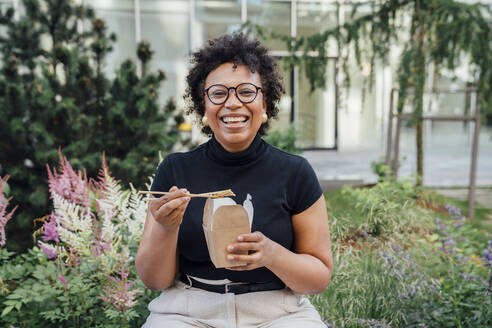 Happy businesswoman wearing eyeglasses sitting with take out food at office park - VPIF06974