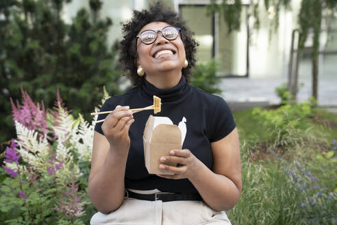 Happy young businesswoman with chopsticks and take out food sitting in office park - VPIF06975