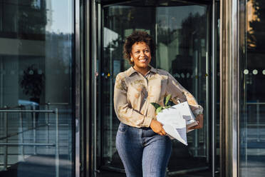 Glückliche junge Geschäftsfrau, die eine Schachtel und Dokumente in der Hand hält und durch eine Tür geht - VPIF07005